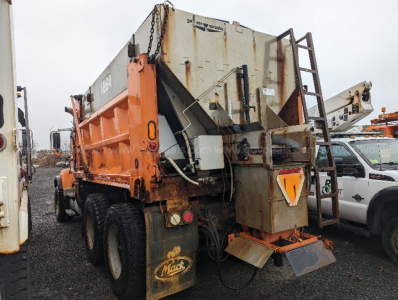 Orange and gray Mack CV713 Granite dump truck viewed from rear left showing plow equipment and warning triangle on back.