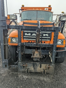 Front view of an orange Mack CV713 Granite plow truck with a heavy-frame front mounting and snow plow attachment; visible text shows 'MACK' on the grille.
