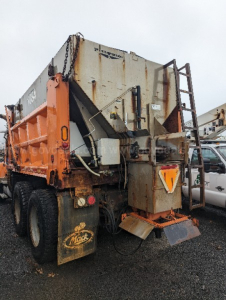 Truck rear view showing plow truck bed with orange rear bumper and Mack mudflap in gravel lot.