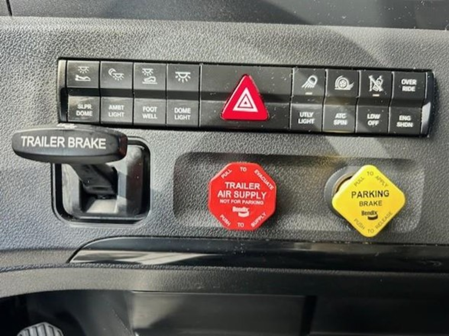 Truck cab dash area with controls: red hazard button, trailer air supply red knob, parking brake yellow knob, and various switches above. Visible text includes TRAILER BRAKE on handle, TRAILER AIR SUPPLY NOT FOR PARKING, PARKING BRAKE, and 