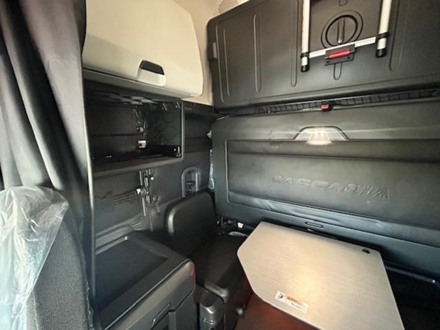Interior of a Freightliner Cascadia truck cab showing side storage shelves, sleeper area, and bulkhead with 'CASCADIA' embossed; compact table folded down and dashboard panels.