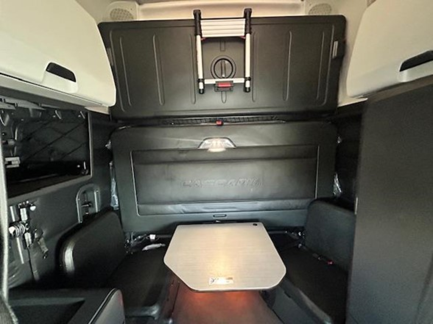 Interior of a Freightliner Cascadia sleeper cab showing a small table and storage compartments, rear wall with a panel, and seating area surrounding the table.