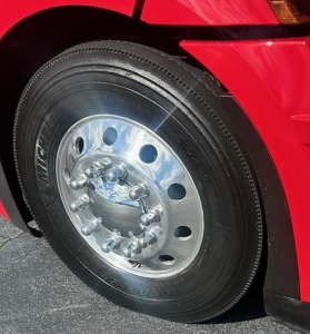 Front wheel of a red Freightliner Cascadia 126 with polished chrome hub and lug nuts, tire shows Invicta branding, fender and body panel in view.