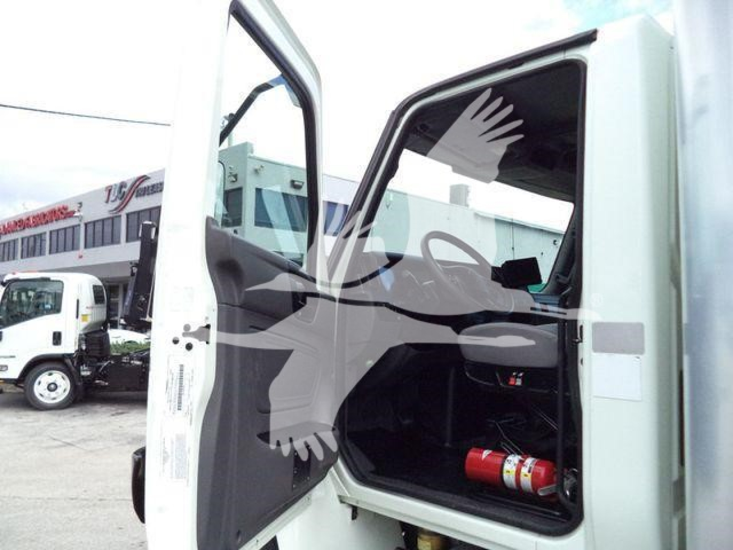 Truck cab interior visible through open door showing driver seat, dashboard, and a red fire extinguisher under bench in cargo area. Side mirrors and exterior white cabin visible.
