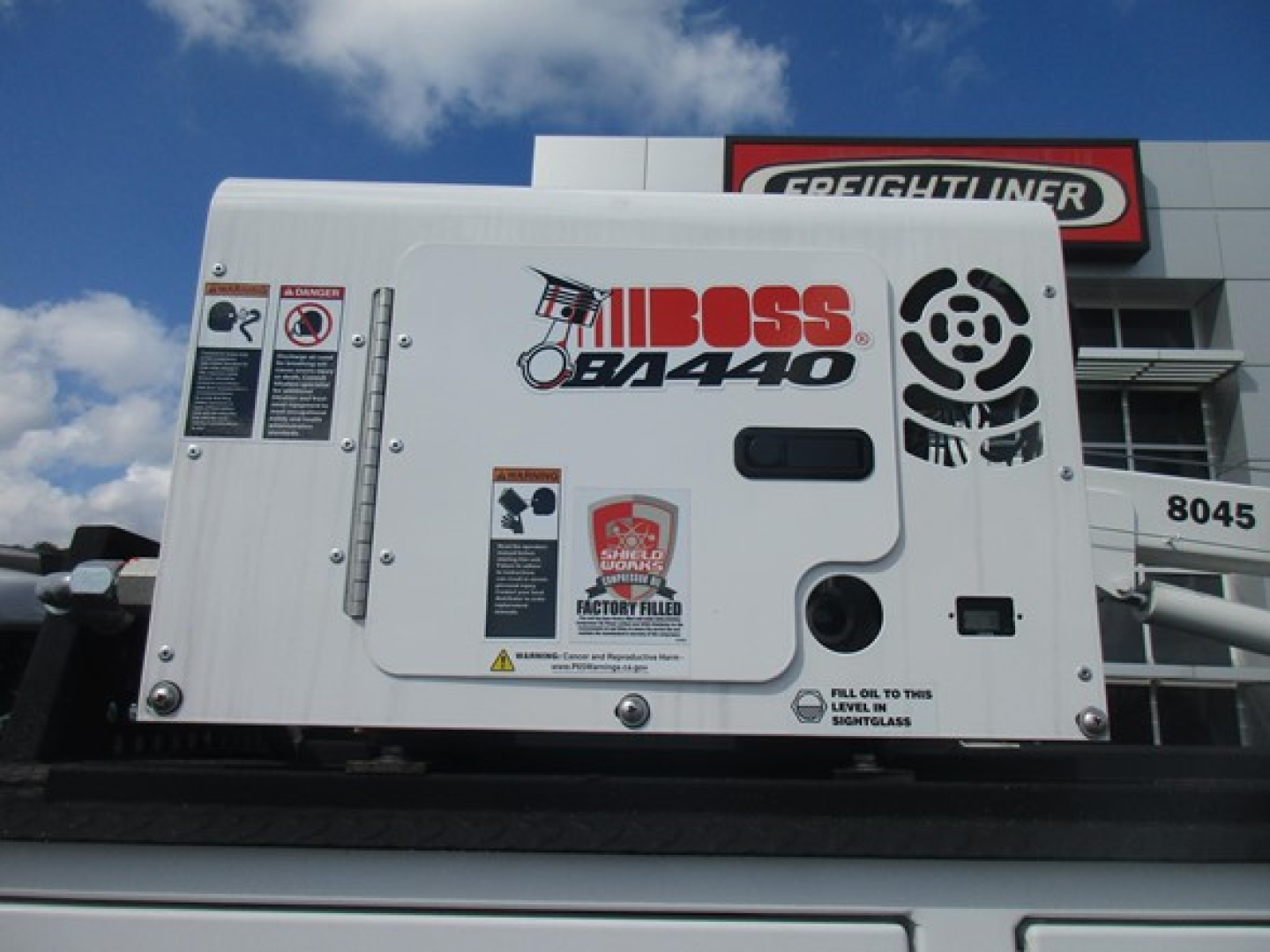 Ford F550 service body truck with Boss 8x4.40 generator unit on top; visible decals include Boss BD4400 and warning labels on the equipment housing; Freightliner dealership in background.
