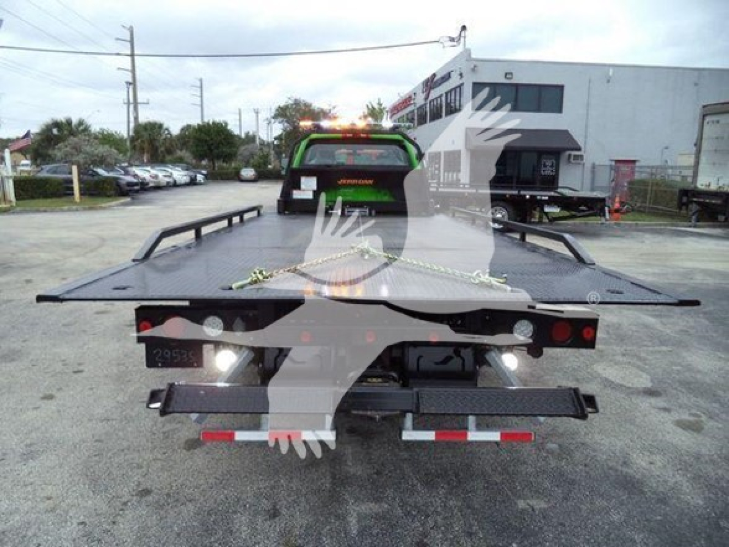Tow truck bed with green-black equipment at rear, flatbed and winch visible, tail lights and bumper, industrial lot in background.
