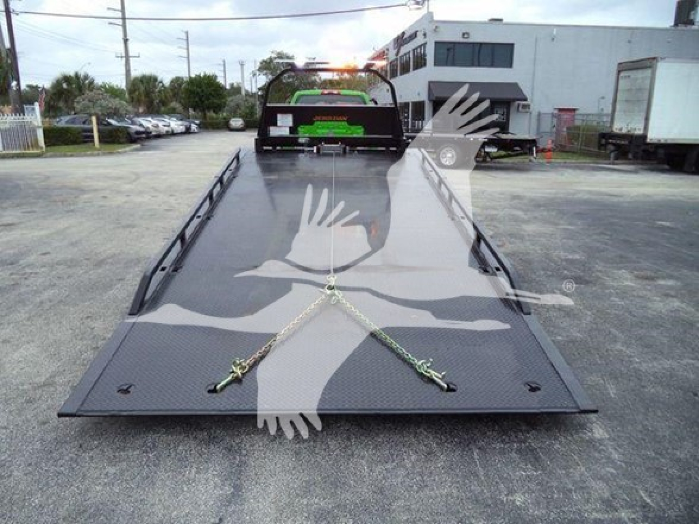 Truck bed with a flat, foldable steel ramp or deck in a truck yard; features embossed diamond plate surface and tie-down chains; background includes a service building and parked vehicles.