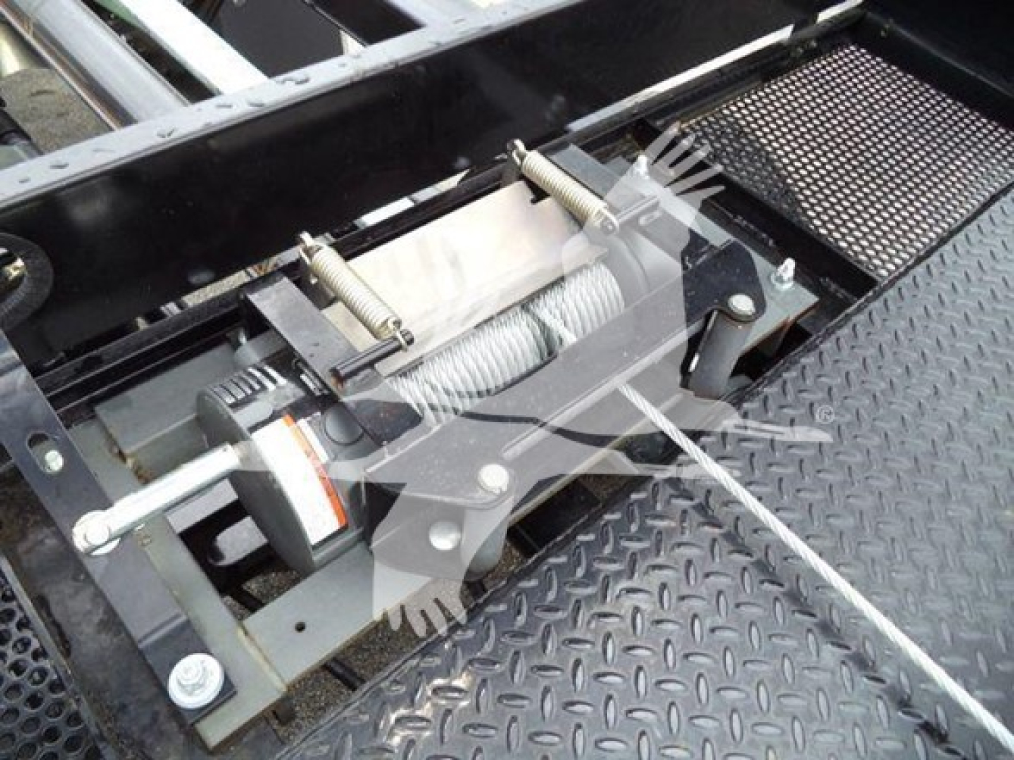 Winch or hoist mechanism on a vehicle rollback bed with steel cable, springs, and mounting brackets visible.