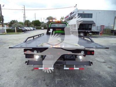 Flatbed tow truck with green-roofed cab seen from rear, loading platform extended, safety chains, rear taillights and reflective strips visible.