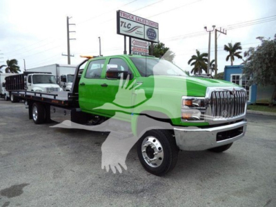Green and white International rollback truck in a lot, front grille chrome, long flatbed, with a watermark graphic across the image. Visible text on signs in background: TLC Truck Sales Center, and utility poles. No legible model or VIN tex