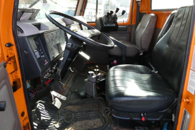 Interior cab of an orange 1999 International dump truck showing black vinyl driver seat, steering wheel, pedals, and dashboard with basic controls.