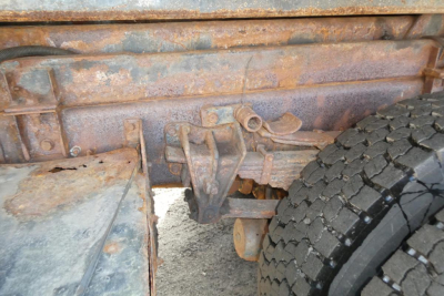 Vehicle chassis showing riveted frame and wheel, with rust and brown dirt, hooklift truck undercarriage and suspension components visible.