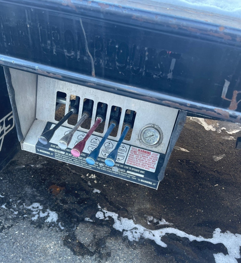 Truck chassis panel with multiple color-coded levers and labels; metal control panel; warning label and gauge visible.