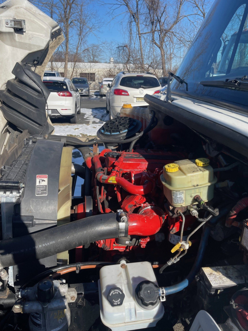 Engine bay of a Freightliner M2 106 medium duty tow truck showing red engine, yellow coolant reservoir, black hoses, and multiple fluid reservoirs with caps.