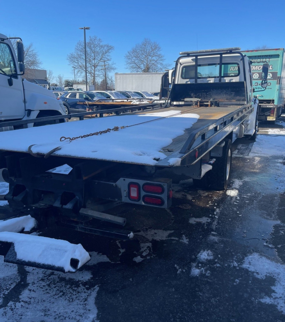 Flatbed tow truck rear and deck showing metal flatbed with snow, taillights, and winch chain visible.