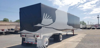 Flatbed trailer with curtain side showing Mac logo and blue/gray cover, rear view, in a lot.