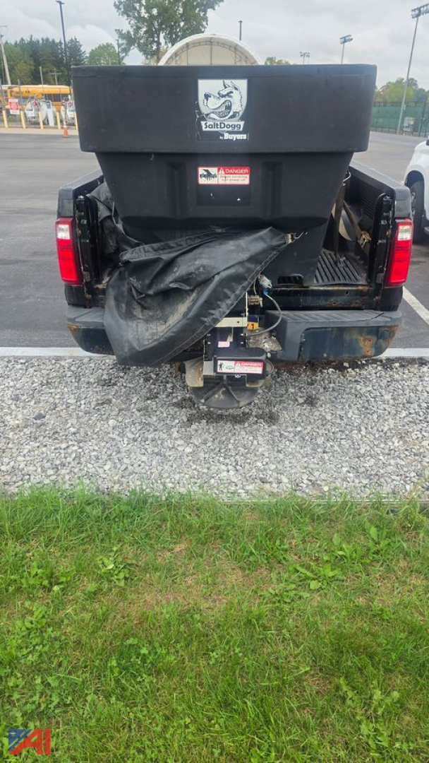 Pickup truck rear view showing salt spreader and plow equipment mounted, with tailgate, bumper, hitch, and debris cover visible.