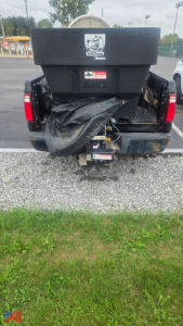 Pickup truck rear view showing salt spreader and plow equipment mounted, with tailgate, bumper, hitch, and debris cover visible.