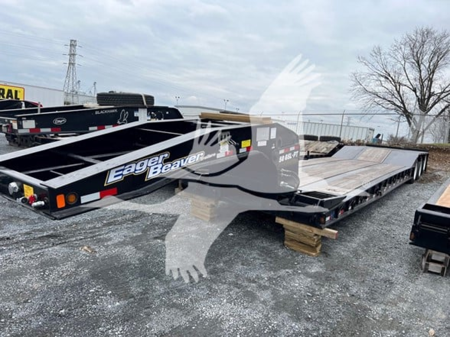 Trailer showing Eager Beaver lowboy tilt deck with 'Eager Beaver' branding and black frame, parked on gravel with wooden supports.