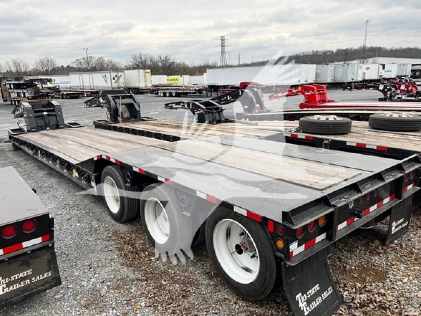 Trailer deck of a lowboy trailer showing flatbed with wood planks and dual rear axles, black frame, wheels, and red and white reflective markings.