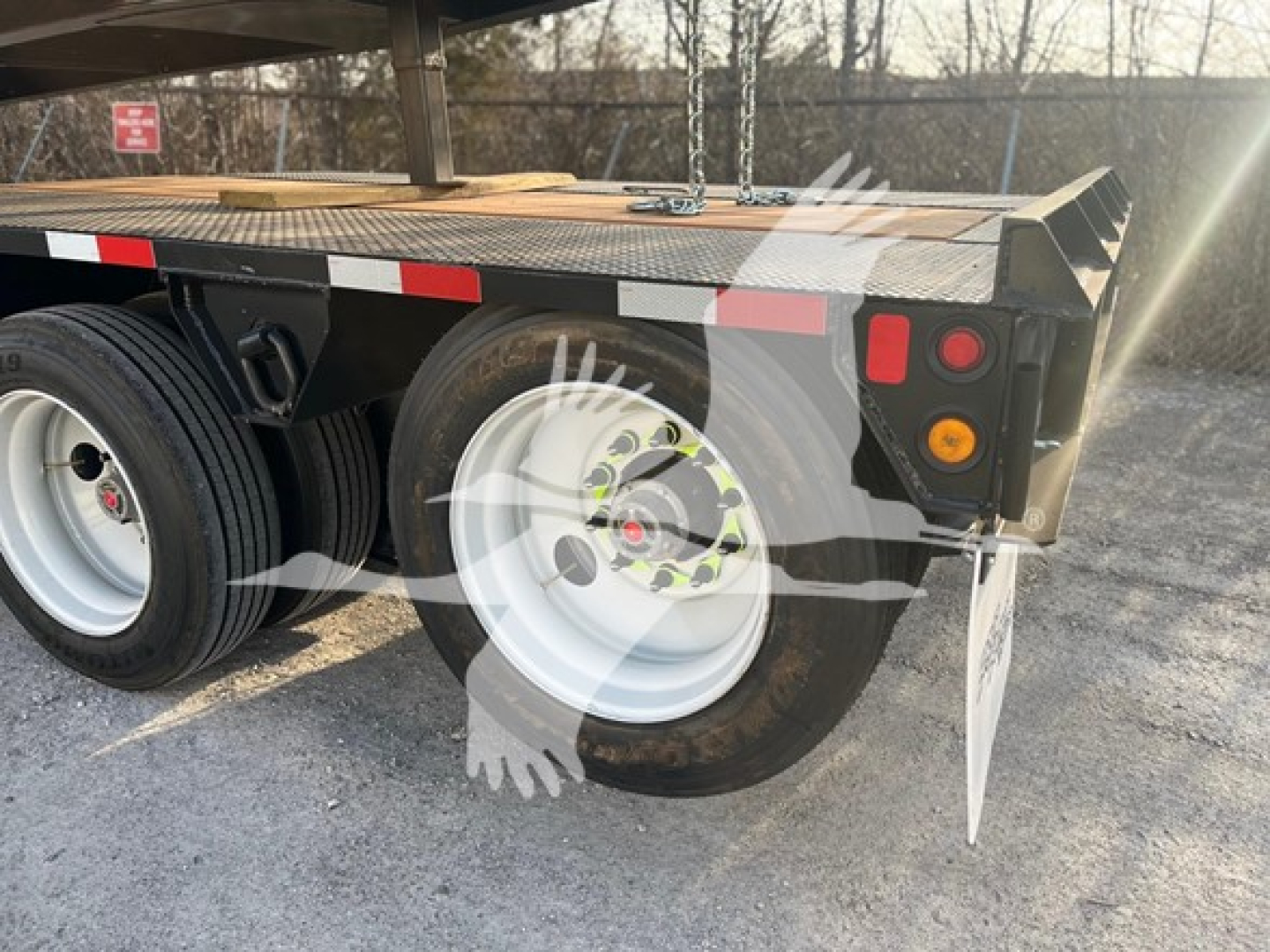 Trailer rear view showing dual rear wheels on a flatbed lowboy trailer with white rims, black fenders, and reflective red/white tape along the edge.