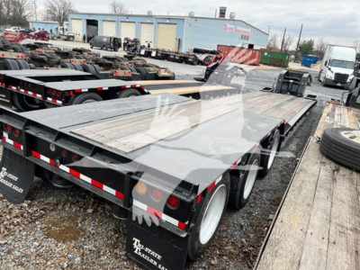 Lowboy trailer showing dual rear axles and flatbed iron with reflective tape and wheel fenders. Visible text includes White Trailer tags and equipment in background.
