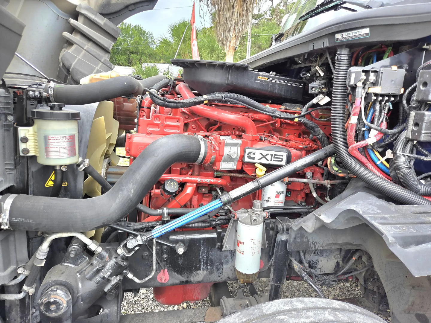Truck engine bay showing Cummins/Isx-15 label on red engine with hoses, filters, and wiring harnesses visible.