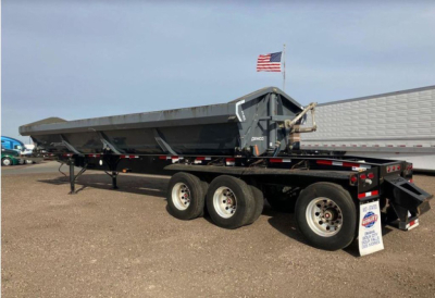 Demco side dump trailer in side view showing gray dump bed, dual-axle tandem wheels, rear ramps, and flag on top of cab.