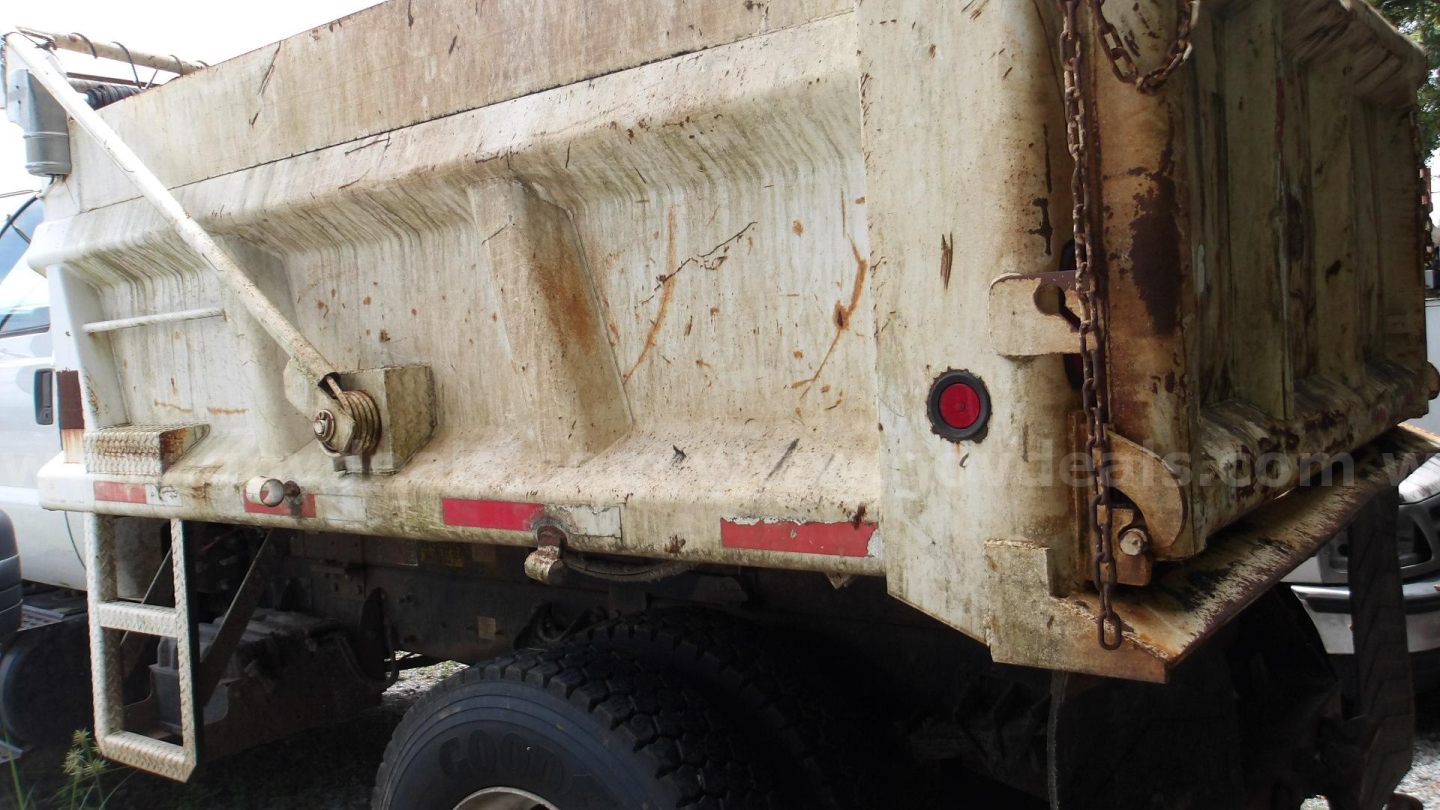 Truck bed of a dump truck with dirt and grime; tailgate latch and rear lights visible; side ladder on left; chain and hooks on rear; weathered metal.