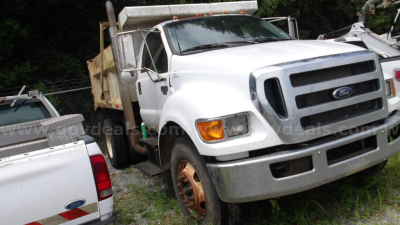 Ford F-750 dump truck front view showing grille, headlights, bumper; visible wheel with rust and side mirror. Text on truck not legible.
