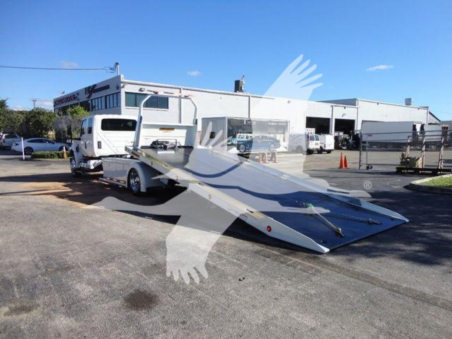 Truck with a flatbed tow ramp attached at the rear, International MV truck in background, dealership lot.
