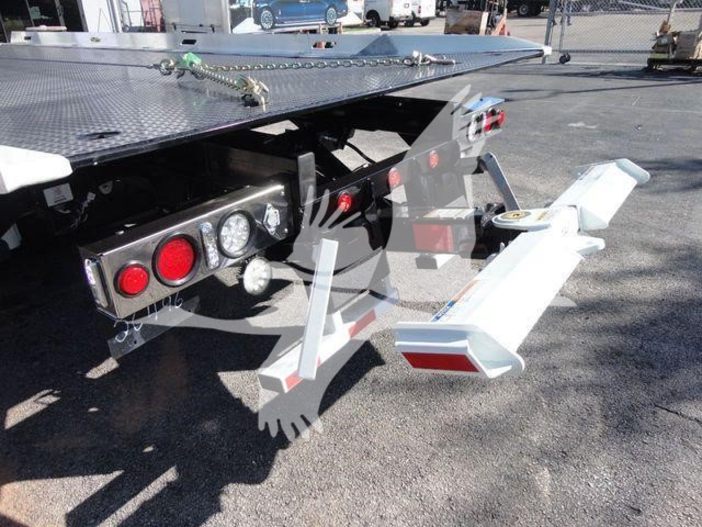 Flatbed truck rear with bumper and hitch assembly showing dual round taillights, red reflectors, and metal support arm extending; platform edge and hydraulic components visible.