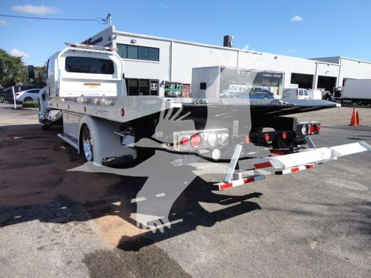Flatbed tow truck in service bay showing long mechanical bed and rear loading ramps with safety lights and reflective tape.