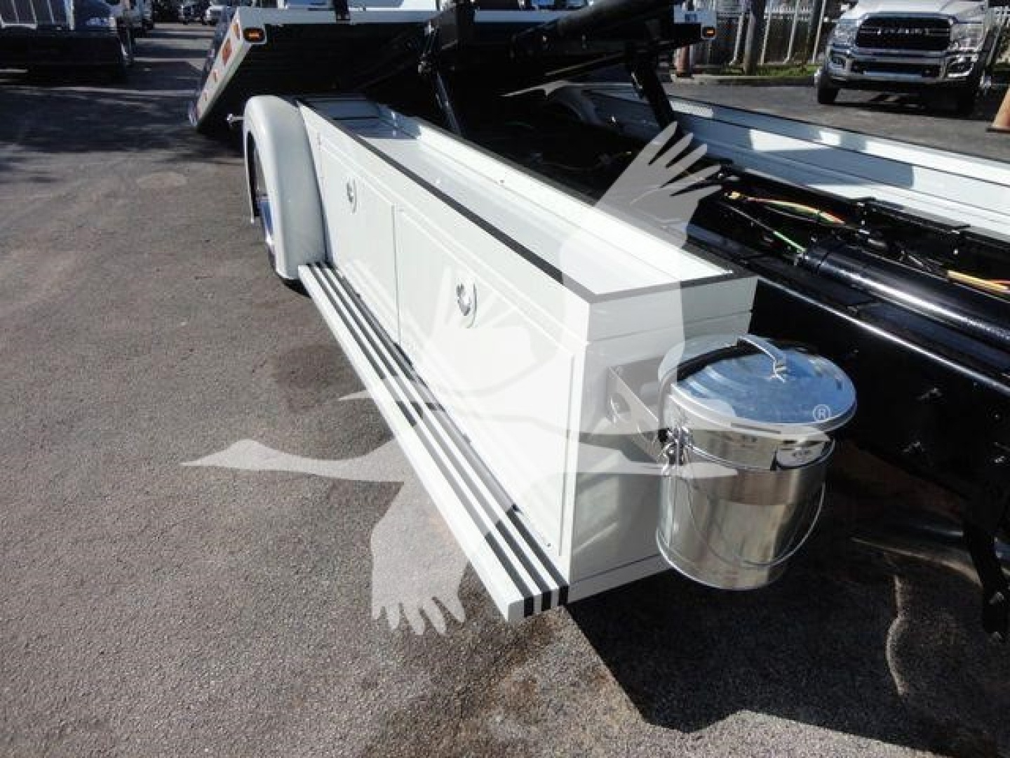 White utility truck bed with side storage drawers and a round chrome fuel or fluid container mounted at the rear; visible hydraulic components and a loading ramp in an outdoor lot.