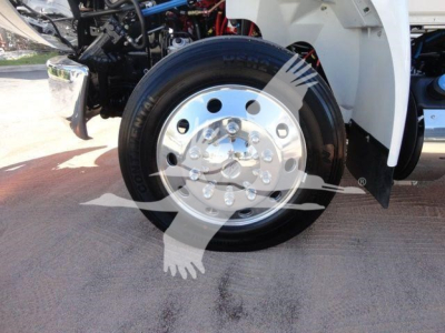 Truck front wheel with shiny chrome hubcap and black tire, engine bay visible behind, part of International MV 2027 in showroom setting.