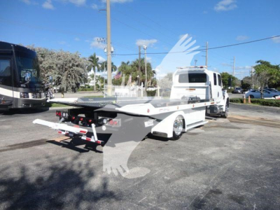 Truck with a white rollback flatbed trailer in a parking lot, front cab visible, other vehicles in background.