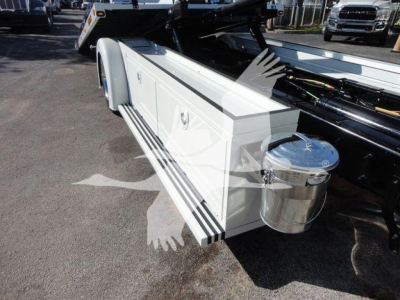 White utility truck bed with side storage drawers and a round chrome fuel or fluid container mounted at the rear; visible hydraulic components and a loading ramp in an outdoor lot.