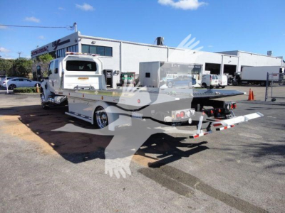 Truck with rollback tow bed in service bay area. Visible text on rear includes company branding and license area, tail lights, and equipment mounts.