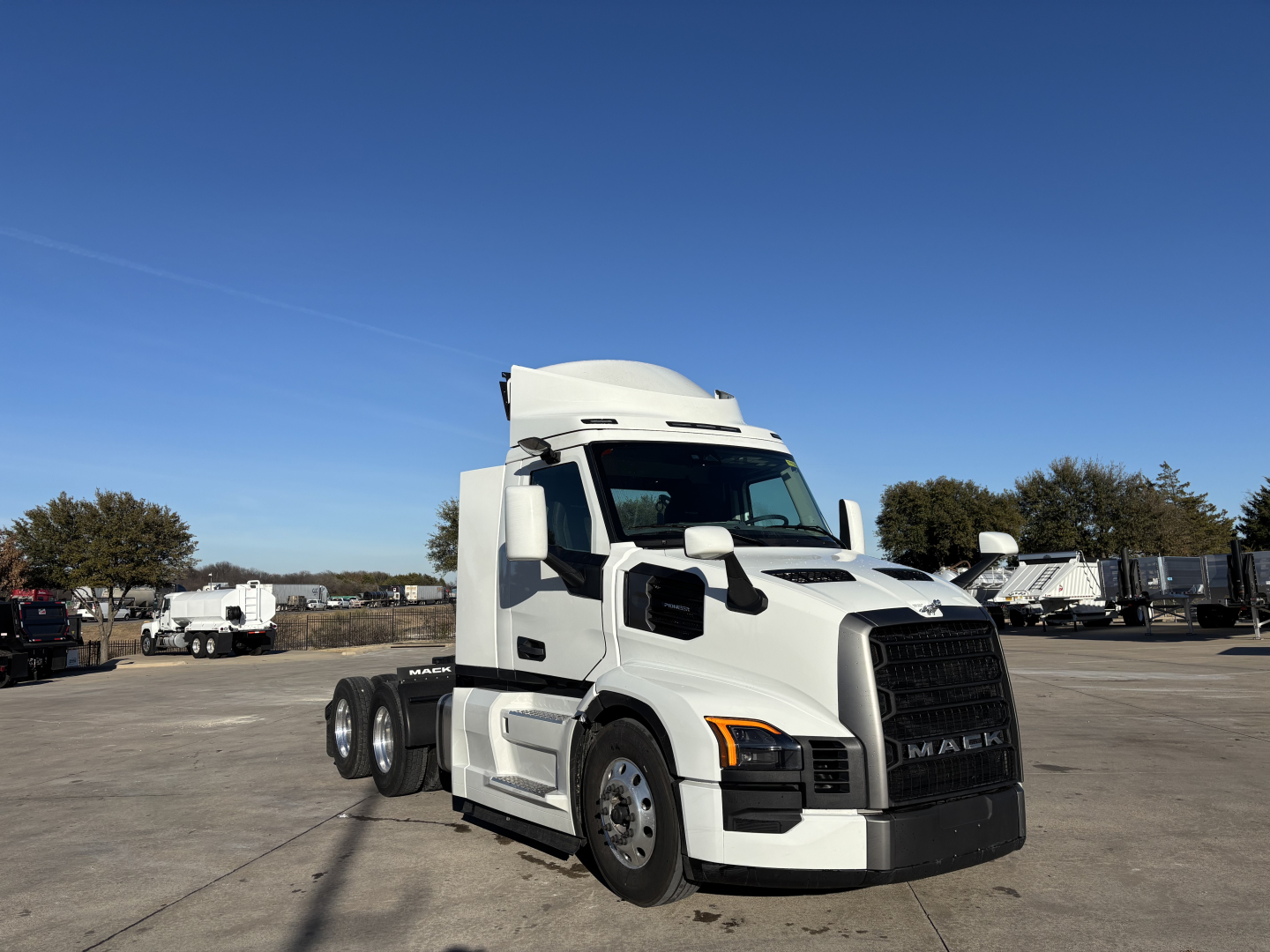 Truck tractor with sleeper cab, white Mack PR64T, in a dealership lot. Front grille with Mack logo, large side mirrors, dual rear axles, black front bumper. Clear blue sky.