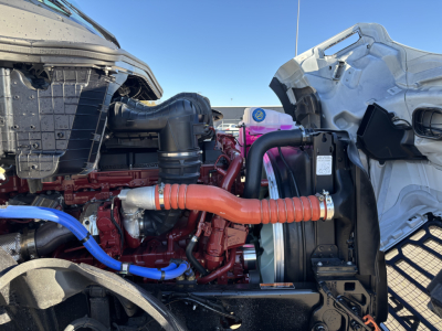 Mack PR64T engine bay showing red engine block, orange high-temperature hose, blue hose, radiator, and intercooler piping visible in a side-by-side cab configuration.