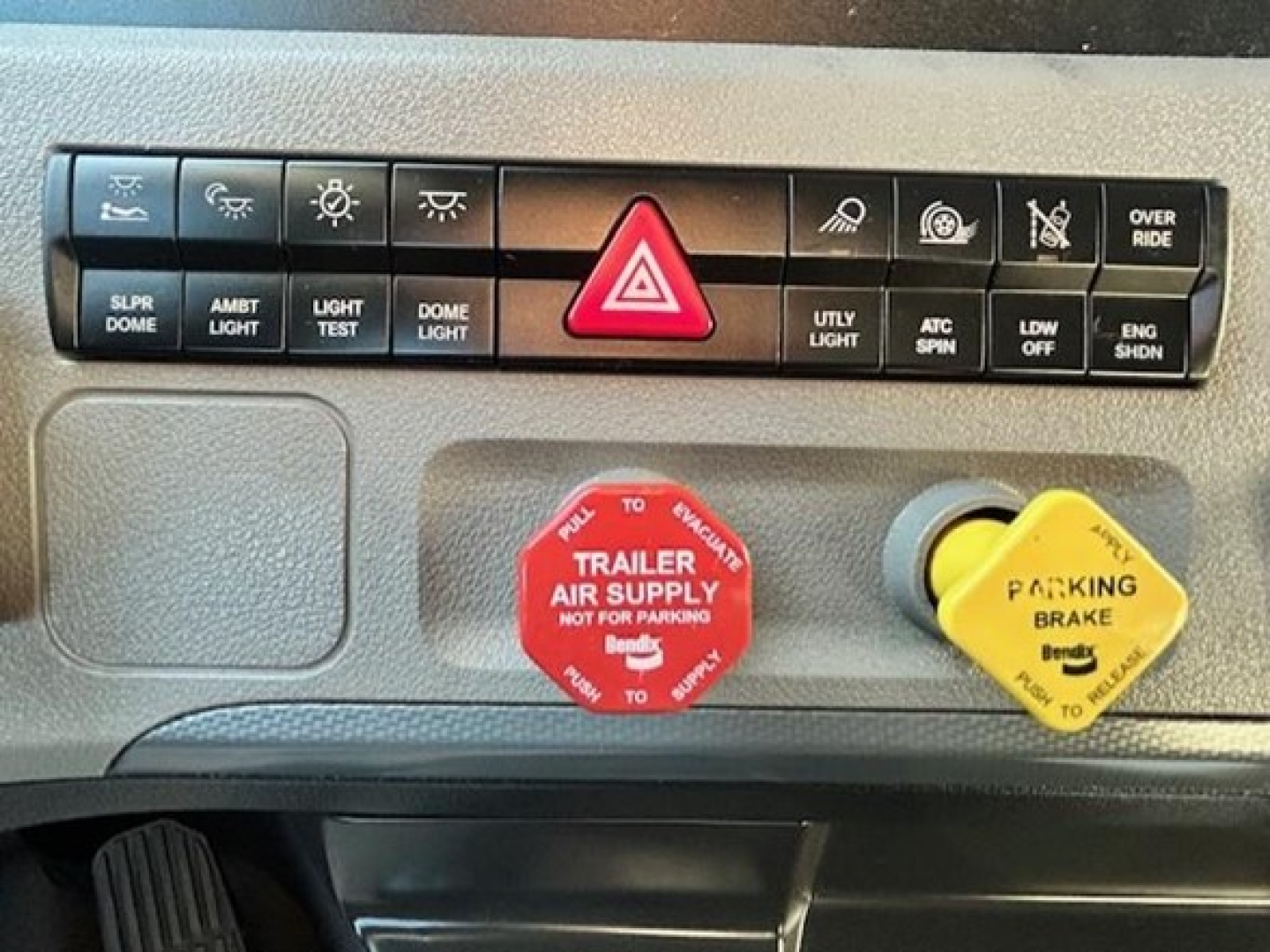 Dashboard switches and controls of a Freightliner Cascadia truck: row of indicator buttons including SLPR DOME, AMBT LIGHT, LIGHT TEST, DOME LIGHT, HAZARD BUTTON, UTILITY LIGHT, ACT SPIN, LDW OFF, ENG SHDN; red trailer air supply cap and ye