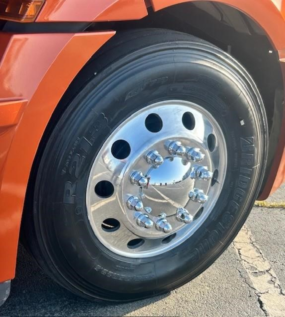Truck wheel on orange Freightliner Cascadia showing polished steel rim with multiple lug nuts and tire.