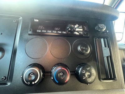 Dashboard controls of a Freightliner Cascadia cabin showing radio unit, three circular dials, and climate controls; visible text includes FM1 frequency display and power outlet label.