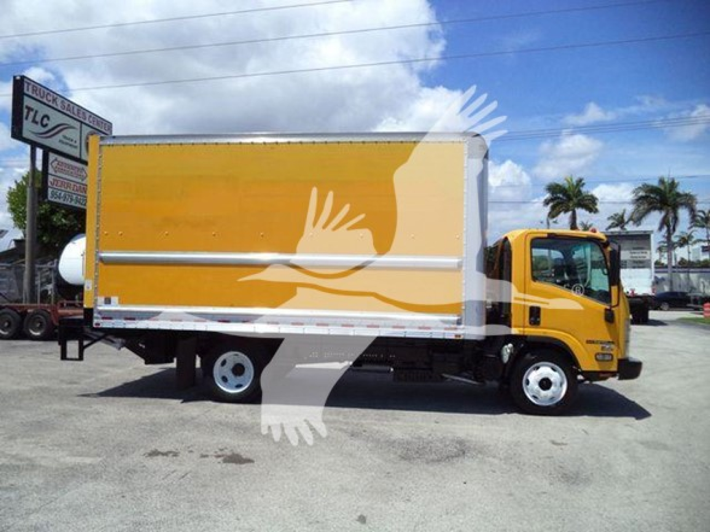 Isuzu NPR-HD box truck with yellow cab and orange cargo box in a lot; visible text on the truck includes no legible labels; background shows palm trees and a dealership sign.