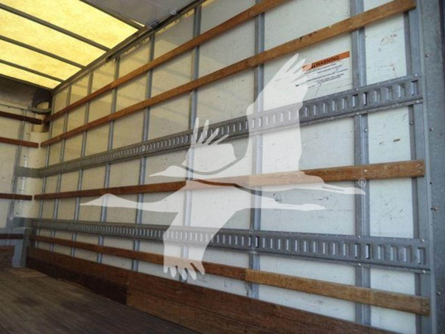Truck box interior showing side wall with vertical metal rails and wooden runners, warning label on wall, empty cargo space.