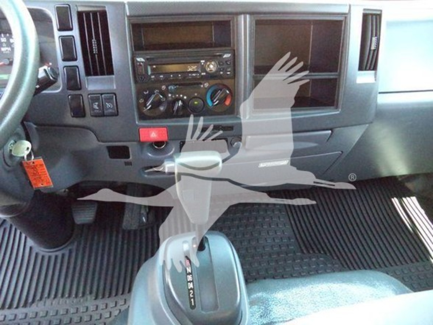 Isuzu NPR HD dashboard and gear selector view showing steering column, center console with radio, climate controls, gear shift labeled P R N D 2 1, and rubber floor mats.