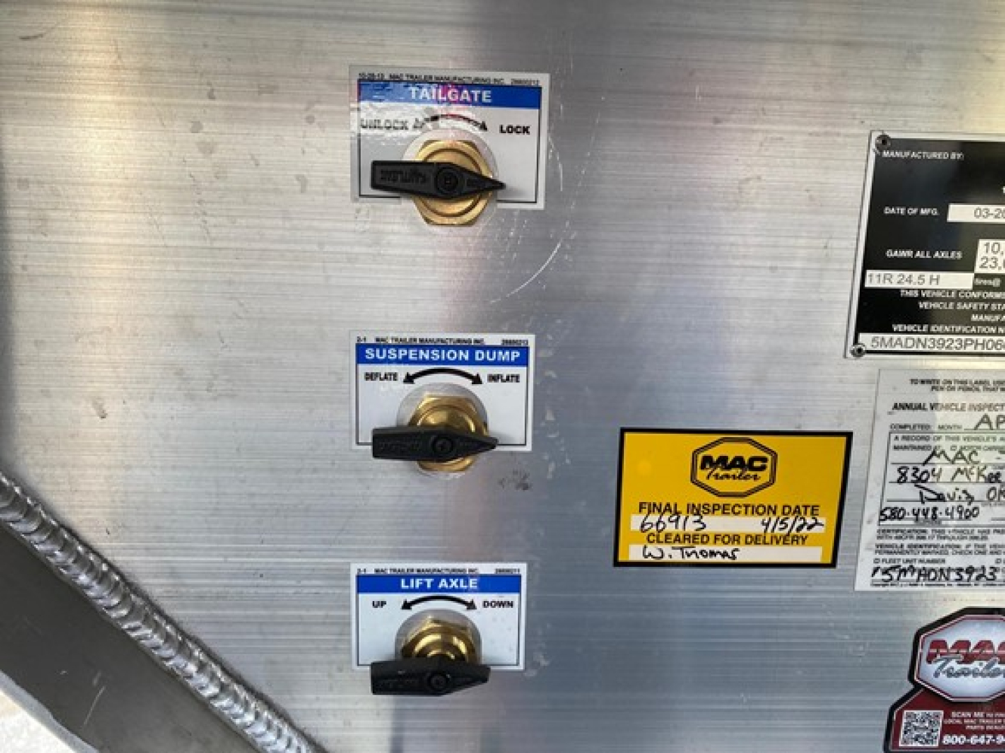 Trailer sidewall with three labeled knobs: tailgate lock, suspension dump, lift axle, and MAC trailer inspection sticker; metal surface with rivets and welded seams.