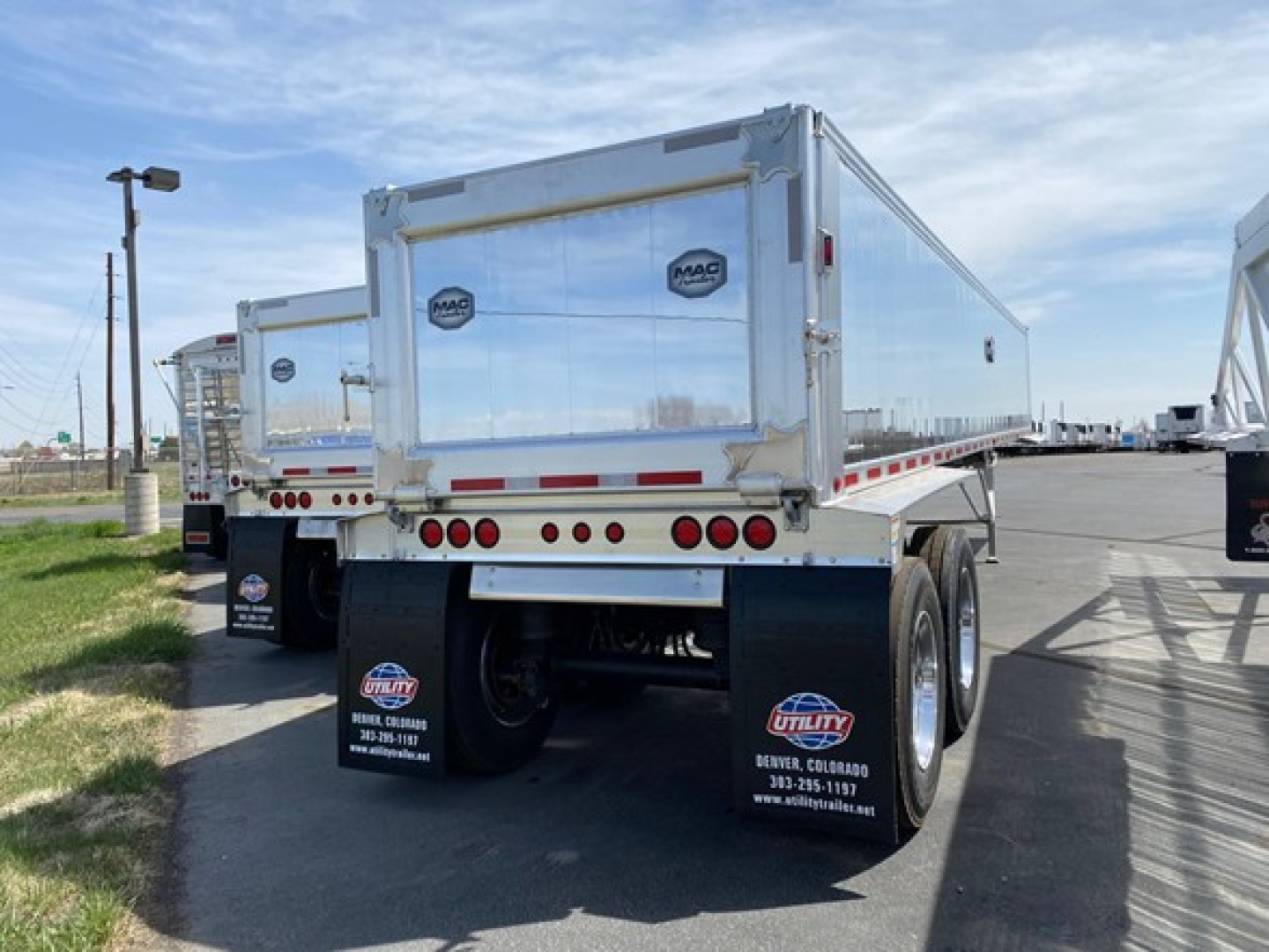 Dump trailer rear view showing MAC aluminum end with MAC logo and dual rear wheels; red reflective tape along lower edge; mud flaps with contact info; clear blue sky.