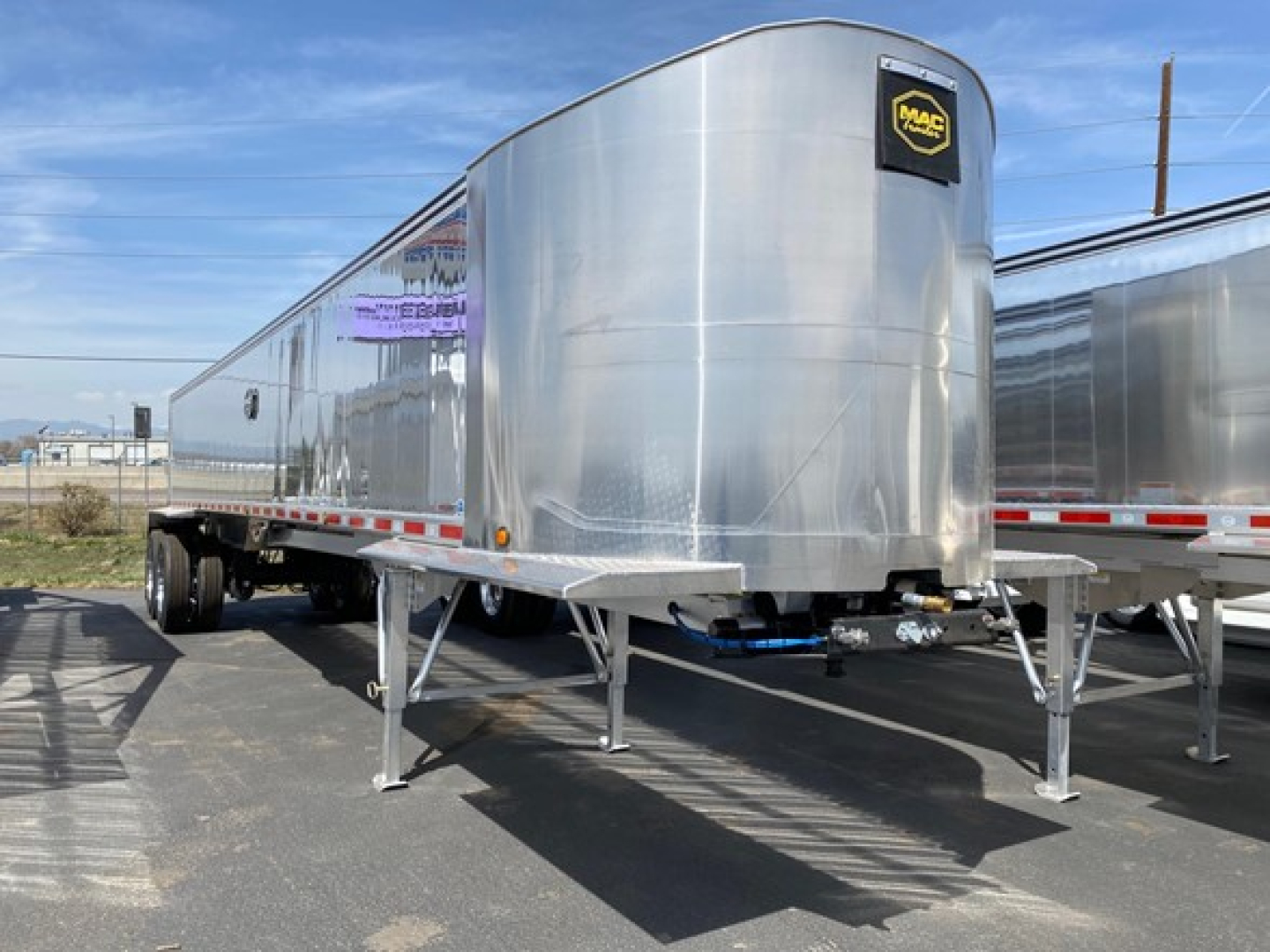 Mac Wes trailer in a lot, polished aluminum dump trailer with blue hitch hoses and Mac logo on front corner.
