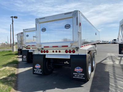 Dump trailer rear view showing MAC aluminum end with MAC logo and dual rear wheels; red reflective tape along lower edge; mud flaps with contact info; clear blue sky.